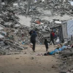 فلسطينيون نازحون يسيرون بين أنقاض المباني المدمرة في حي الشيخ رضوان (الأوروبية)